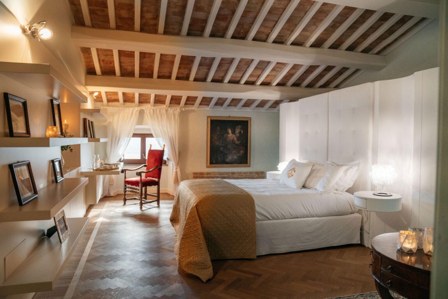 Spacious and elegantly designed bedroom featuring a high wooden ceiling, a plush bed with decorative pillows, a cozy armchair, and tasteful artwork. Natural light filters through sheer curtains, highlighting the warm tones of the hardwood floor and modern furnishings. Ideal for relaxation and comfort. | Only on SopranoVillas
