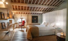 Spacious and elegantly designed bedroom featuring a high wooden ceiling, a plush bed with decorative pillows, a cozy armchair, and tasteful artwork. Natural light filters through sheer curtains, highlighting the warm tones of the hardwood floor and modern furnishings. Ideal for relaxation and comfort.