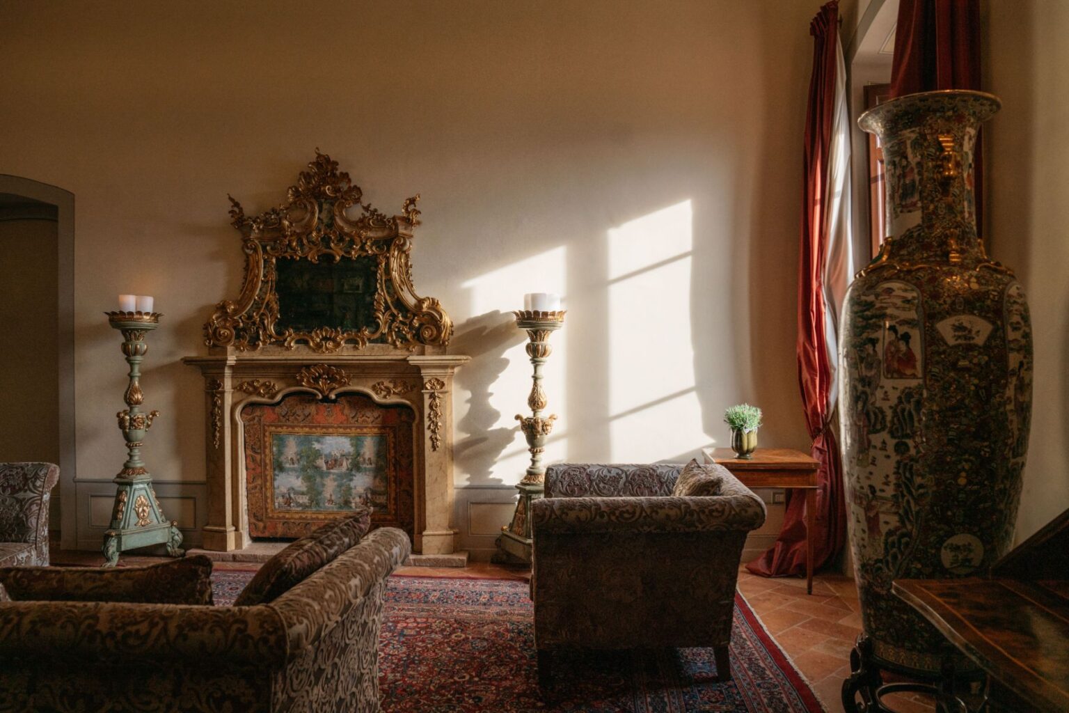 Elegant living room featuring ornate fireplace, vintage armchairs, decorative vases, and natural light streaming through large windows. The space is adorned with rich textiles and a cozy atmosphere, ideal for relaxation and gatherings. | Only on SopranoVillas
