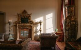 Elegant living room featuring ornate fireplace, vintage armchairs, decorative vases, and natural light streaming through large windows. The space is adorned with rich textiles and a cozy atmosphere, ideal for relaxation and gatherings.