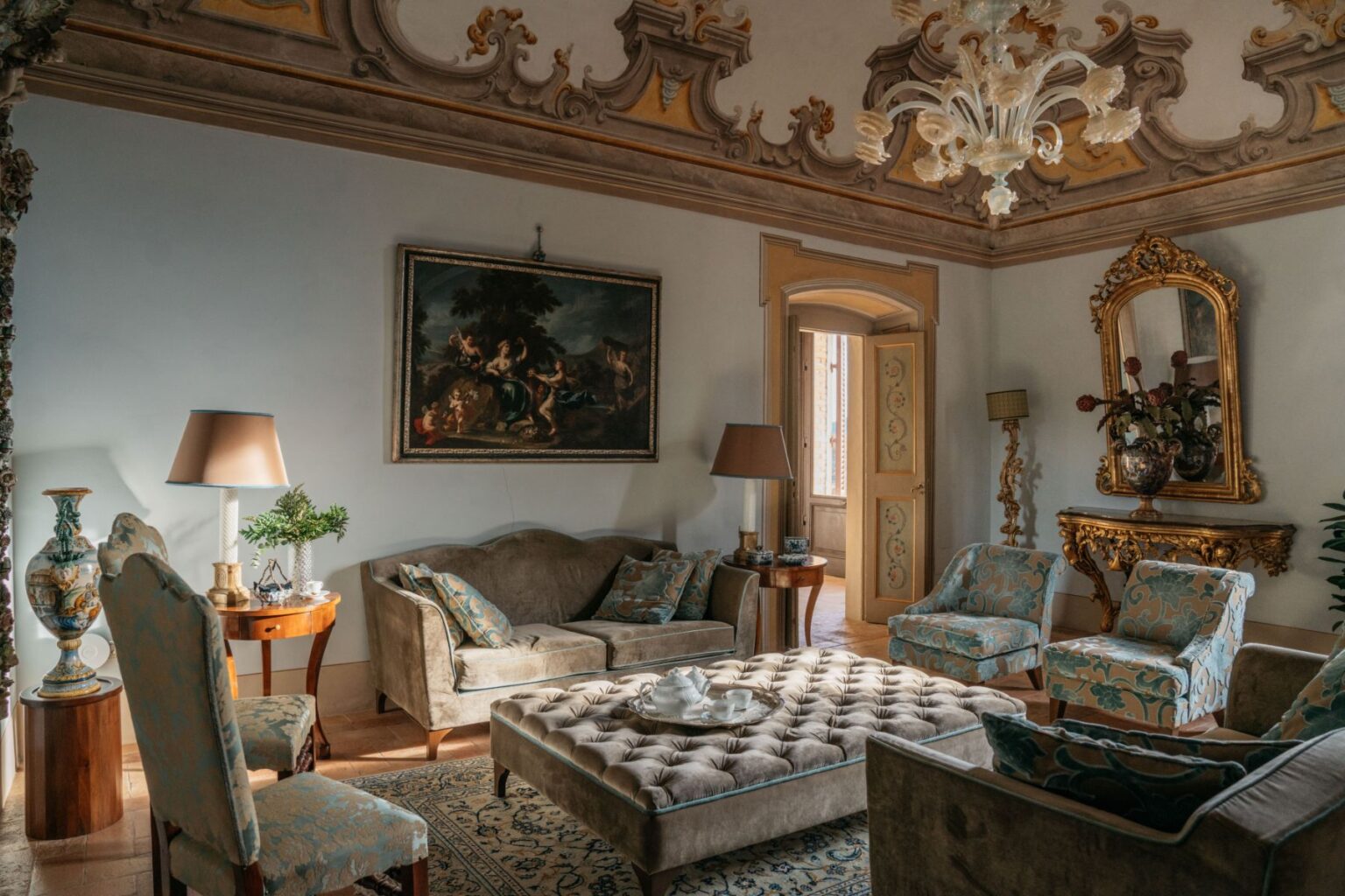 Elegant living room featuring ornate ceiling details, plush furniture with decorative cushions, and a classic painting. The space includes a stylish coffee table, decorative lamp, and a golden-framed mirror, creating a sophisticated atmosphere. Natural light streams in through an open door, enhancing the inviting ambiance. | Only on SopranoVillas