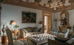 Elegant living room featuring ornate ceiling details, plush furniture with decorative cushions, and a classic painting. The space includes a stylish coffee table, decorative lamp, and a golden-framed mirror, creating a sophisticated atmosphere. Natural light streams in through an open door, enhancing the inviting ambiance.