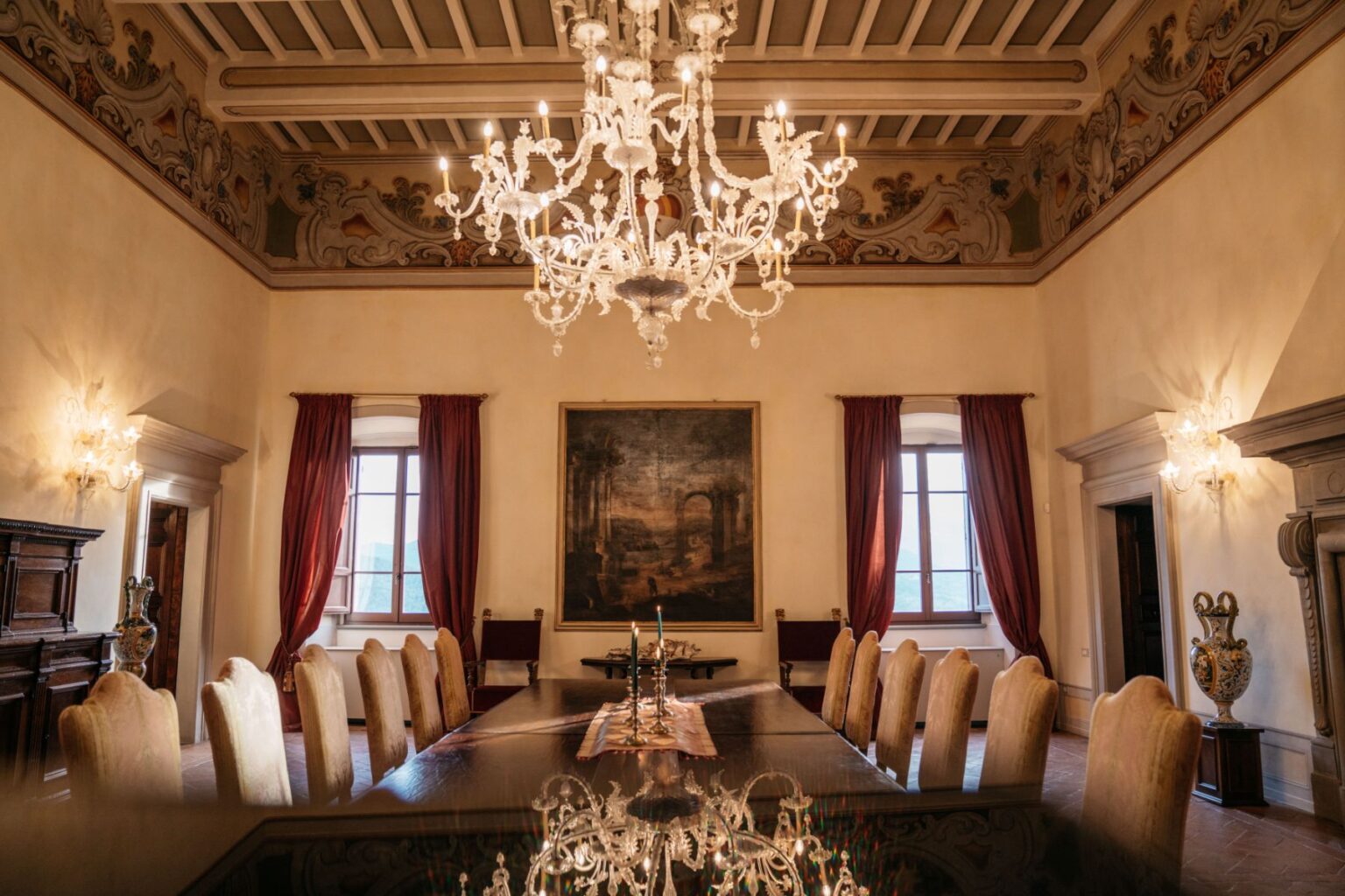 Elegant dining room with a large wooden table surrounded by plush chairs, featuring a stunning chandelier and decorative frescoes on the ceiling. The room is illuminated by natural light from large windows, creating a warm ambiance. A classic painting adorns the wall, complemented by rich red curtains and ornate furnishings. Ideal for formal gatherings and events. | Only on SopranoVillas