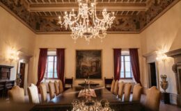 Elegant dining room with a large wooden table surrounded by plush chairs, featuring a stunning chandelier and decorative frescoes on the ceiling. The room is illuminated by natural light from large windows, creating a warm ambiance. A classic painting adorns the wall, complemented by rich red curtains and ornate furnishings. Ideal for formal gatherings and events.