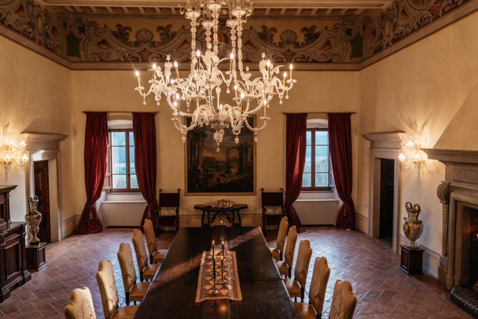 Elegant dining room featuring a long wooden table surrounded by ornate chairs, illuminated by a crystal chandelier. The room is adorned with rich red drapes, decorative wall paintings, and a classic fireplace, creating a warm and inviting atmosphere. | Only on SopranoVillas