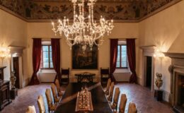 Elegant dining room featuring a long wooden table surrounded by ornate chairs, illuminated by a crystal chandelier. The room is adorned with rich red drapes, decorative wall paintings, and a classic fireplace, creating a warm and inviting atmosphere.