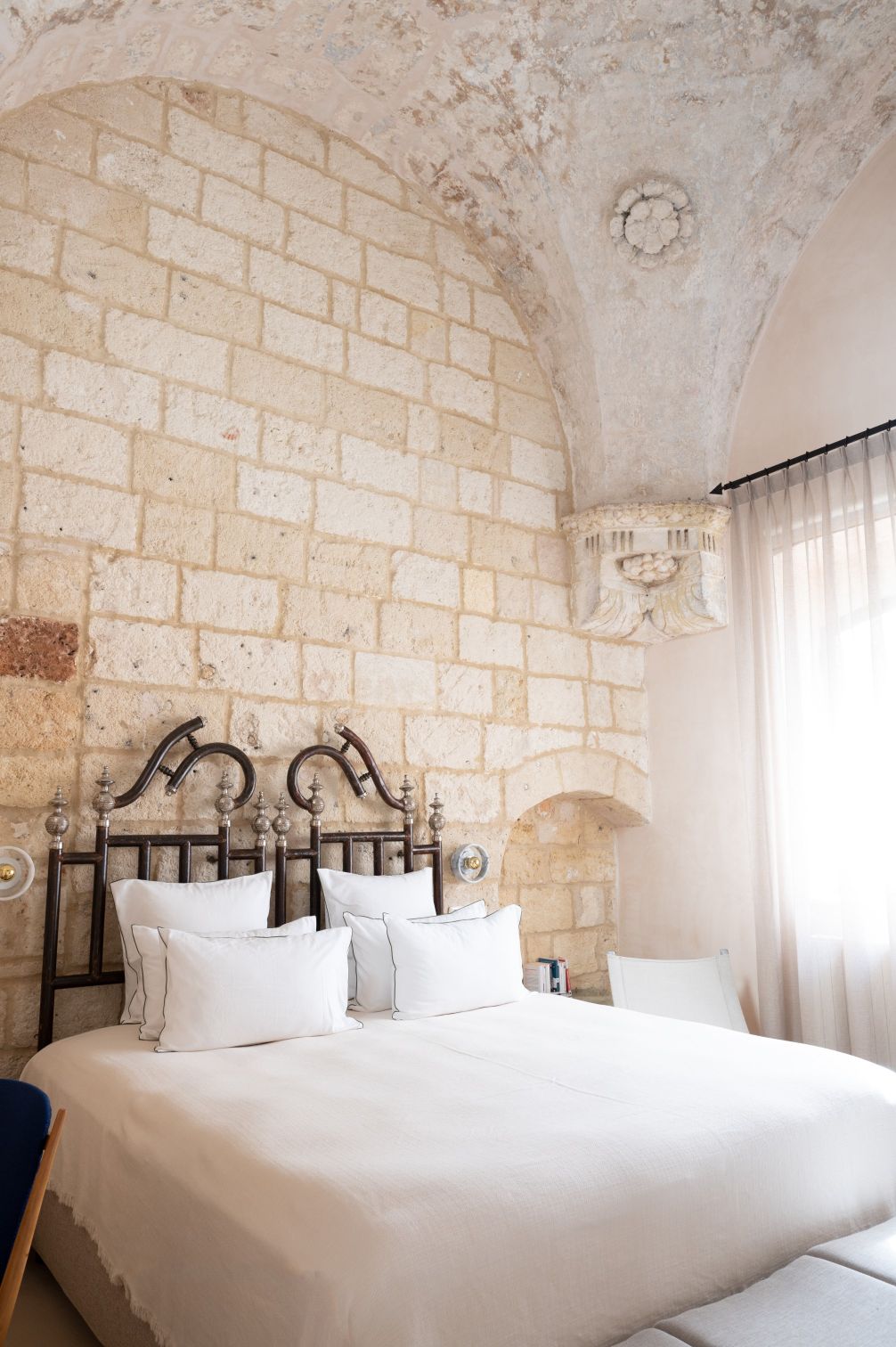 Elegant bedroom featuring a large bed with white linens and decorative pillows, set against a rustic stone wall and arched ceiling. Natural light filters through sheer curtains, enhancing the cozy atmosphere. | Only on SopranoVillas