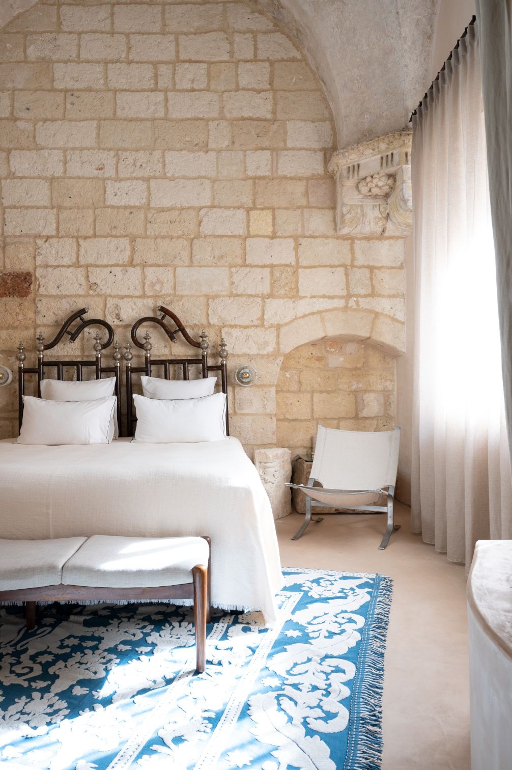 Cozy bedroom featuring a double bed with decorative iron headboards, white linens, and a soft beige throw. The room has exposed stone walls, a patterned blue area rug, a minimalist chair, and sheer curtains allowing natural light to filter in. | Only on SopranoVillas