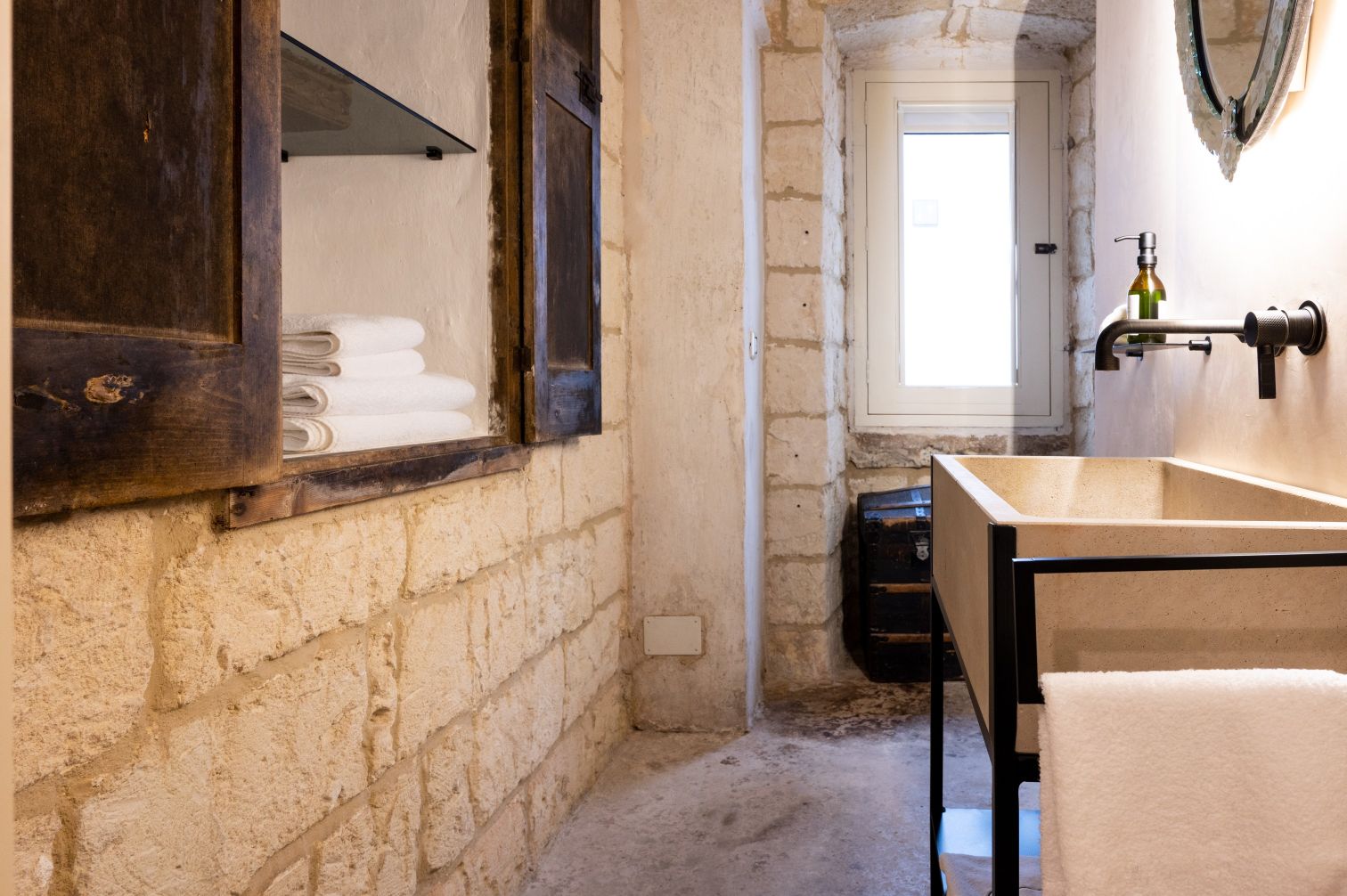 Modern bathroom featuring natural stone walls, a minimalist sink, neatly stacked towels, and a vintage storage trunk. The space is illuminated by natural light from a window, enhancing its rustic charm. | Only on SopranoVillas