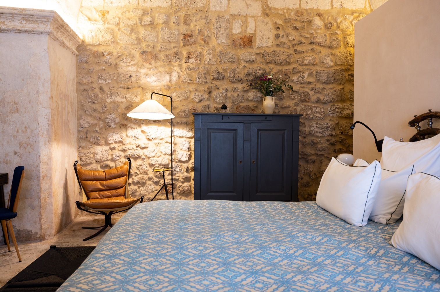Cozy bedroom featuring a blue patterned bedspread, white pillows, and a rustic stone wall. The room includes a stylish brown leather chair, a modern floor lamp, and a dark blue cabinet with decorative flowers on top, creating a warm and inviting atmosphere. | Only on SopranoVillas