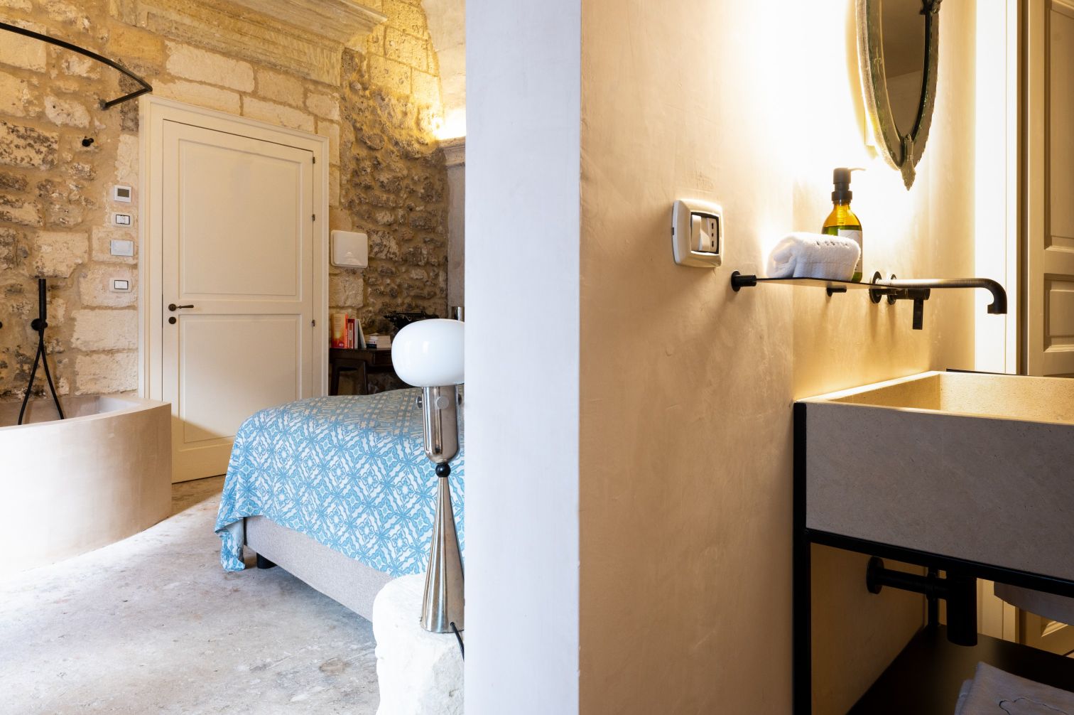 Interior view of a stylish hotel room featuring a cozy bed with a blue patterned cover, a modern bathroom sink with a towel and soap, and exposed stone walls, creating a warm and inviting atmosphere. | Only on SopranoVillas