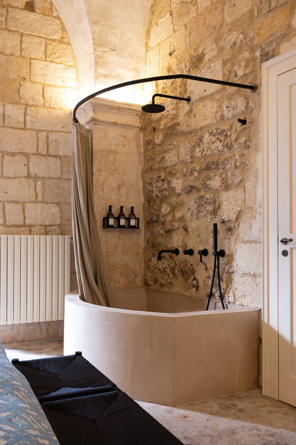Stylish bathroom featuring a modern curved bathtub with a black shower fixture, set against rustic stone walls. The space includes a sleek curtain, a minimalist shelf with three bottles, and a radiator, creating a blend of contemporary and vintage design elements. | Only on SopranoVillas