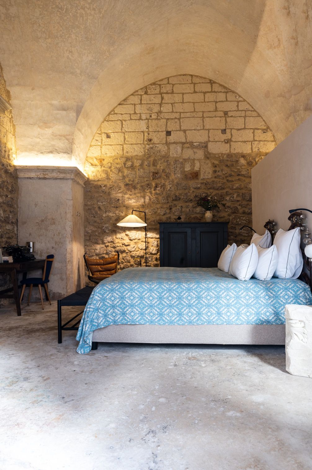 Cozy bedroom interior featuring a blue patterned bedspread, stone walls, and an arched ceiling. The space includes a wooden cabinet, a stylish lamp, and a comfortable armchair, creating a warm and inviting atmosphere. | Only on SopranoVillas