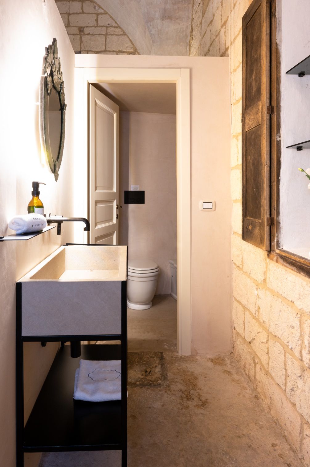 Modern bathroom interior featuring a minimalist sink with a black frame, a wall-mounted mirror, and a wooden cabinet. The space includes a toilet in the background and showcases natural stone walls and flooring, creating a stylish and functional ambiance. | Only on SopranoVillas