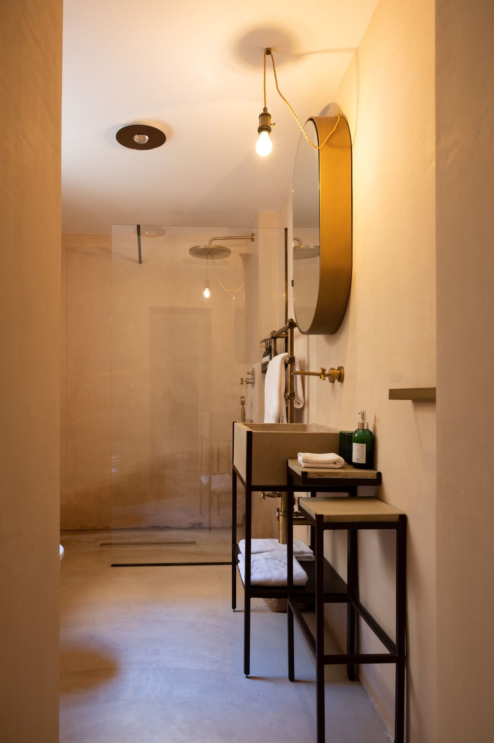 Modern bathroom interior featuring a minimalist design with a glass shower, stylish round mirror, and a sleek sink unit. The space is illuminated by a pendant light and has neutral-toned walls, creating a serene atmosphere. | Only on SopranoVillas