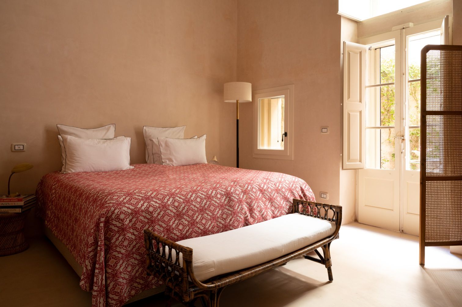 Cozy bedroom featuring a double bed with a red patterned quilt, white pillows, and a stylish wicker bench. Natural light streams in through open French windows, illuminating the warm, neutral-toned walls. A modern lamp and a woven basket add to the room's inviting decor. | Only on SopranoVillas