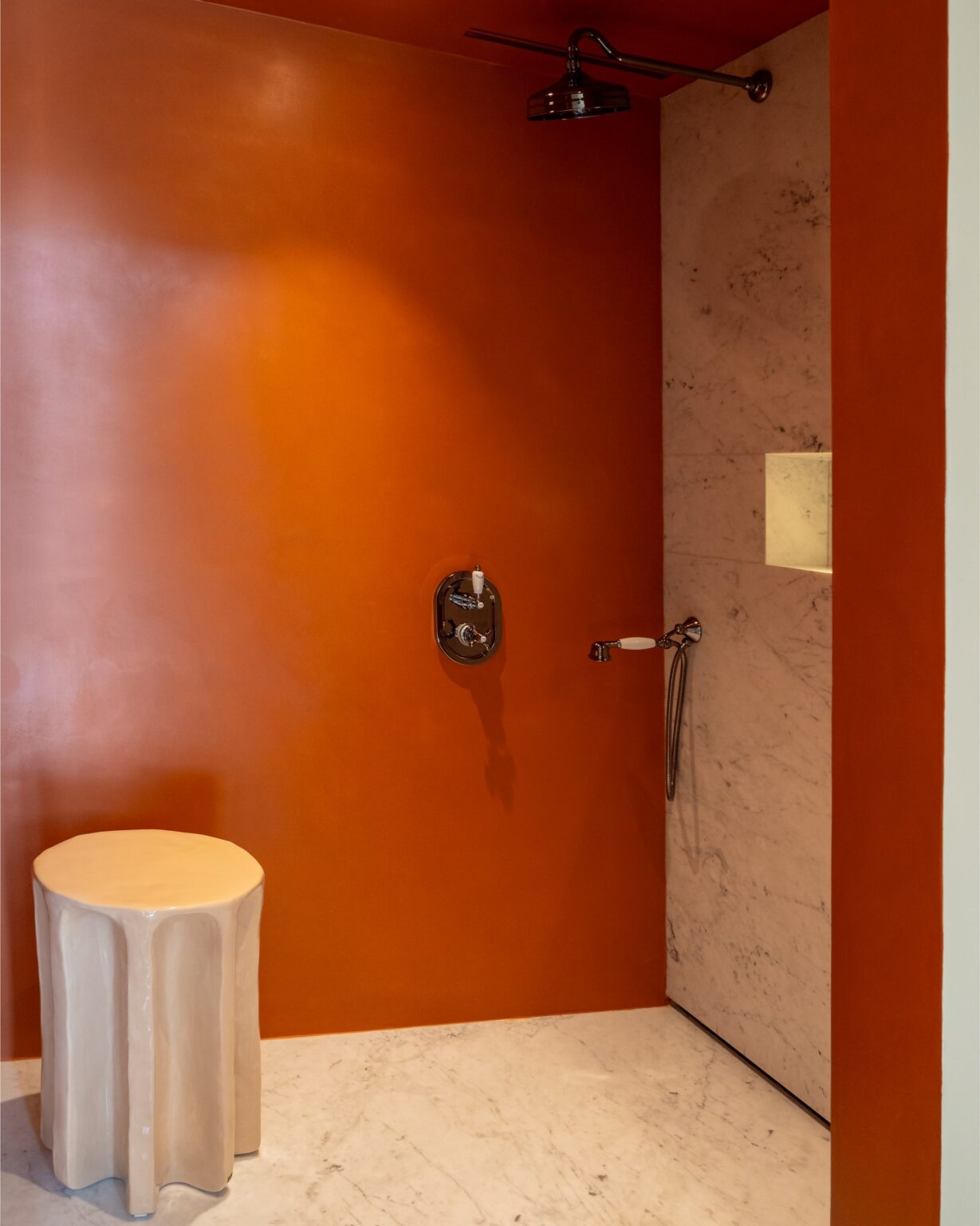 Modern shower space with an orange textured wall, a marble panel, and a stylish showerhead, featuring a unique beige stool for seating. Ideal for contemporary bathroom design inspiration. | Only on SopranoVillas