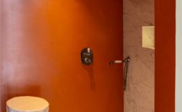 Modern shower space with an orange textured wall, a marble panel, and a stylish showerhead, featuring a unique beige stool for seating. Ideal for contemporary bathroom design inspiration.