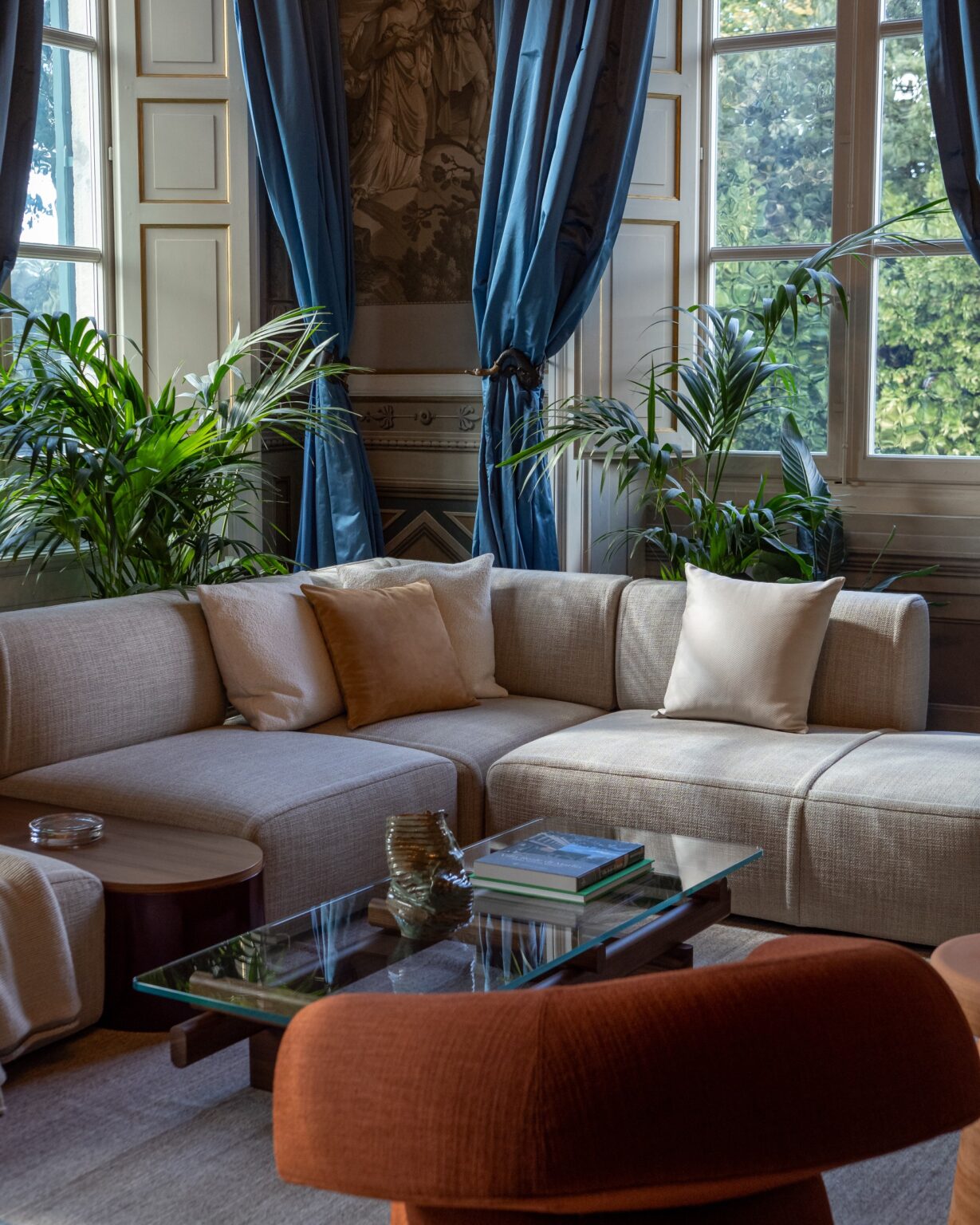 Elegant living room featuring a light-colored sectional sofa with decorative pillows, a glass coffee table with books and a decorative vase, and vibrant green plants. Blue curtains frame large windows, enhancing the room's bright and airy atmosphere. | Only on SopranoVillas