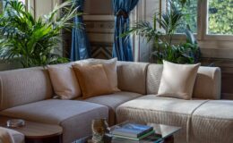 Elegant living room featuring a light-colored sectional sofa with decorative pillows, a glass coffee table with books and a decorative vase, and vibrant green plants. Blue curtains frame large windows, enhancing the room's bright and airy atmosphere.