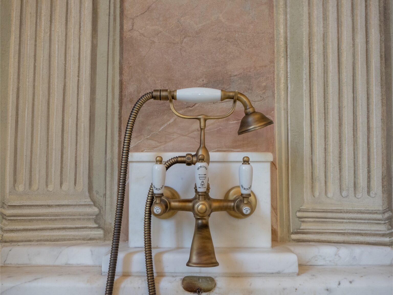 Antique brass faucet with a handheld showerhead, featuring white ceramic handles, mounted on a marble countertop against a textured wall. Ideal for vintage bathroom design inspiration. | Only on SopranoVillas