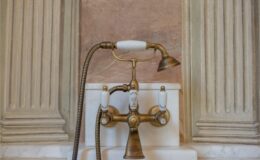 Antique brass faucet with a handheld showerhead, featuring white ceramic handles, mounted on a marble countertop against a textured wall. Ideal for vintage bathroom design inspiration.