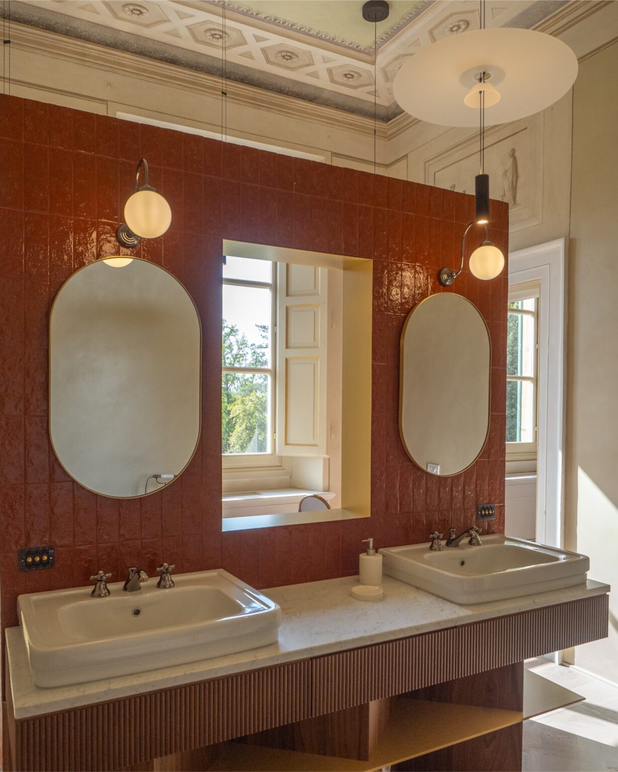 Modern bathroom interior featuring dual sinks, elegant oval mirrors, and stylish pendant lighting against a rich red tiled wall, with a window showcasing a view of greenery outside. | Only on SopranoVillas