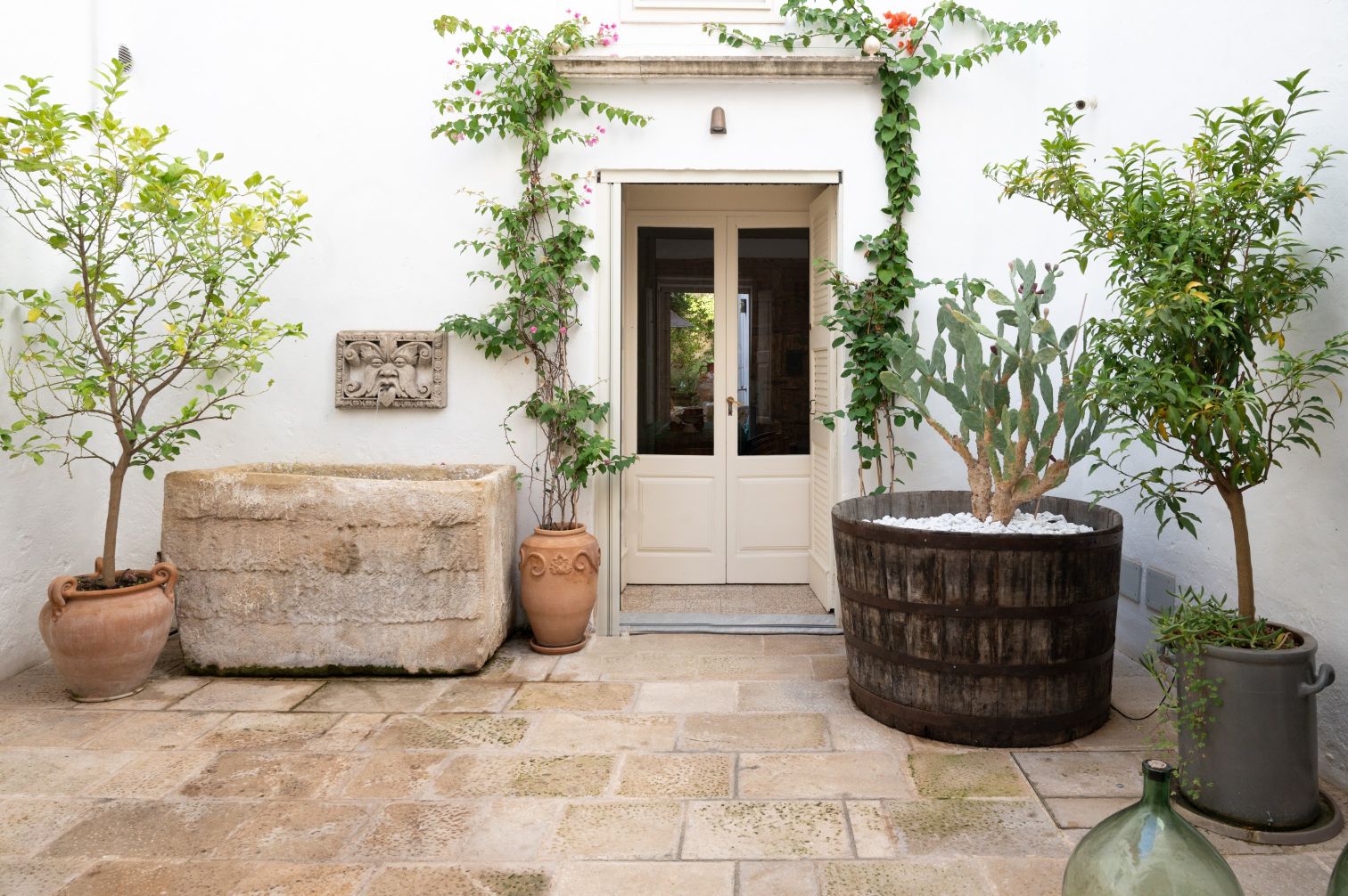 Charming outdoor courtyard featuring a stone water trough, potted plants, and a wooden planter with a cactus, framed by a white wall and a double door entrance. | Only on SopranoVillas