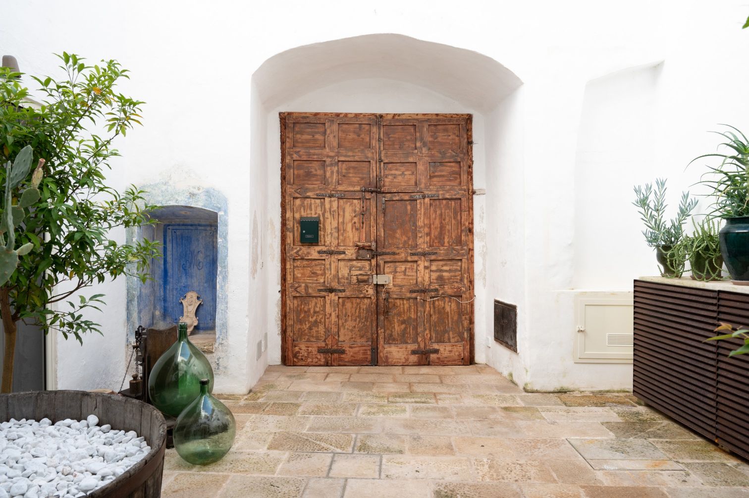 Wooden double doors with rustic detailing set in a whitewashed wall, surrounded by greenery and decorative elements like a blue niche and glass bottles. The scene captures a serene courtyard ambiance, ideal for design inspiration. | Only on SopranoVillas