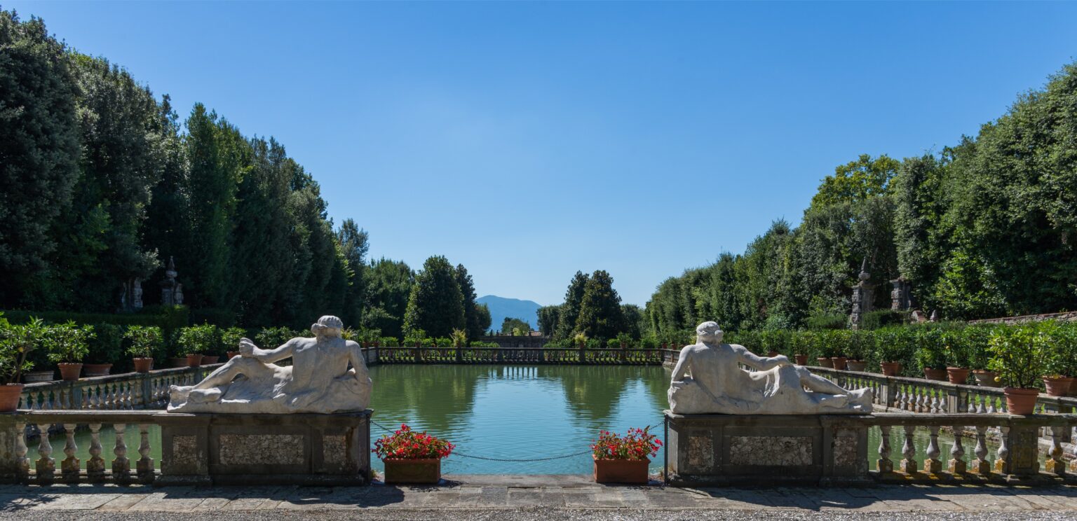 Scenic view of a tranquil garden featuring classical stone sculptures beside a serene pond, surrounded by lush greenery and potted plants, under a clear blue sky. Perfect for outdoor relaxation and historical exploration. | Only on SopranoVillas