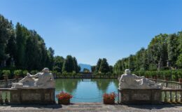 Scenic view of a tranquil garden featuring classical stone sculptures beside a serene pond, surrounded by lush greenery and potted plants, under a clear blue sky. Perfect for outdoor relaxation and historical exploration.