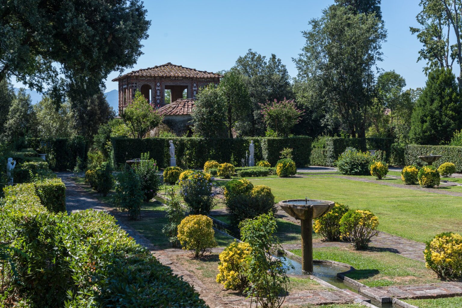 Lush garden landscape featuring neatly trimmed hedges, vibrant yellow and green shrubs, and decorative fountains, with a rustic building in the background under clear blue skies. Ideal for nature enthusiasts and garden lovers. | Only on SopranoVillas
