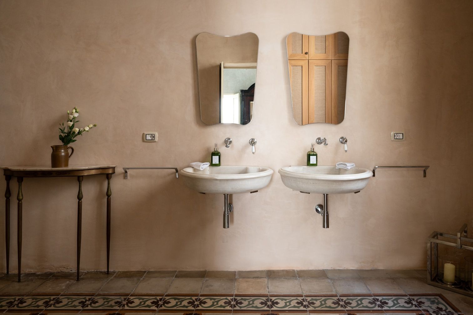 Elegant bathroom design featuring two modern sinks, stylish mirrors, and a vintage table with a vase of flowers. The warm, earthy tones and decorative tiles enhance the inviting atmosphere. | Only on SopranoVillas