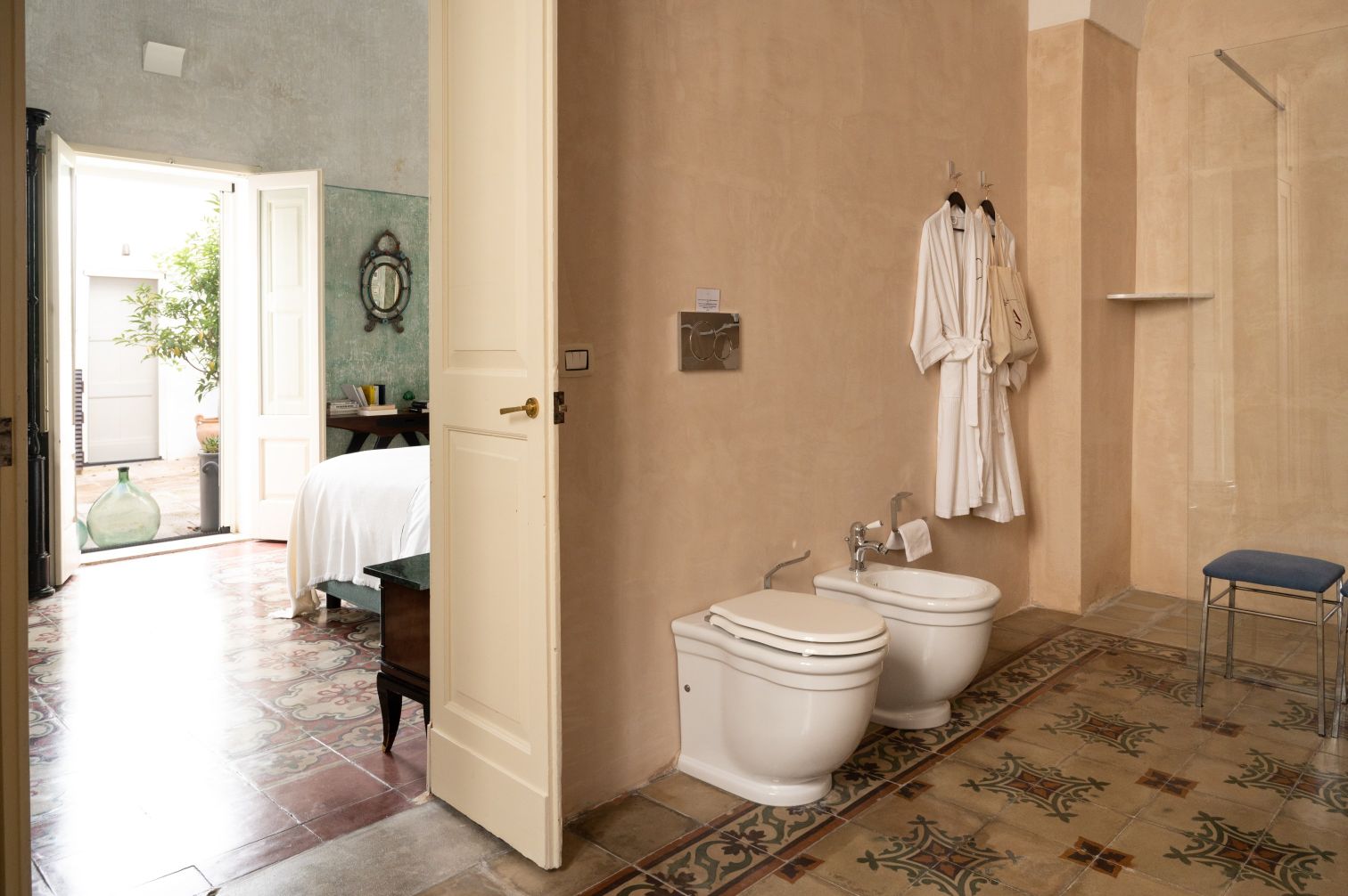 Modern bathroom featuring two white toilets, a glass shower, and hanging bathrobes. The room has a rustic ambiance with patterned tile flooring and an open door leading to a bright outdoor area with greenery. | Only on SopranoVillas