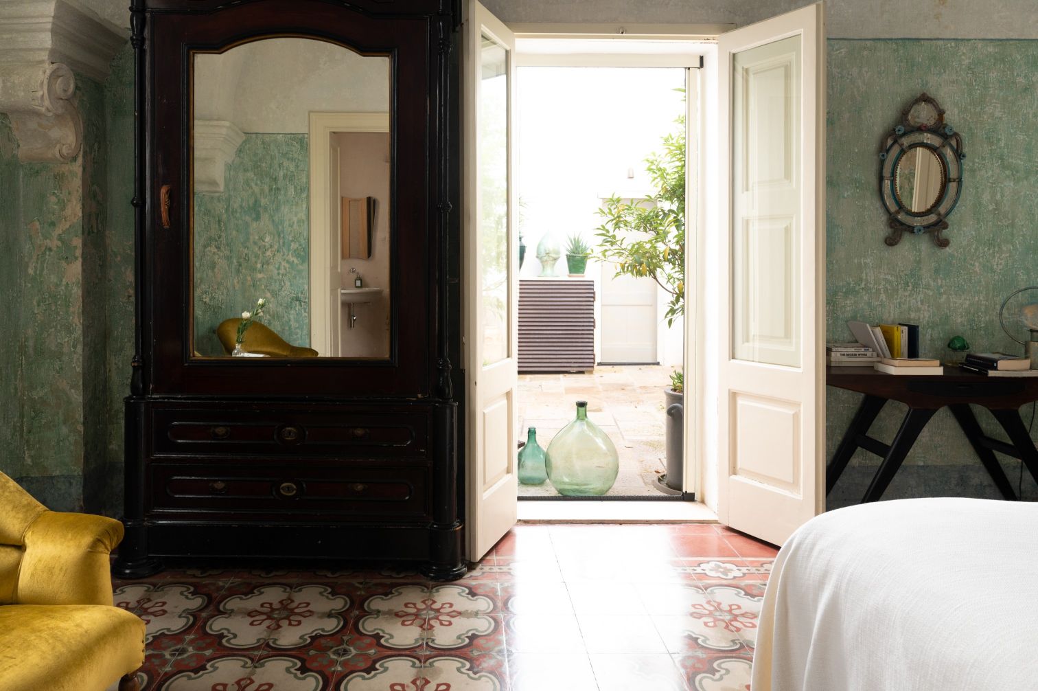 Interior of a stylish bedroom featuring a vintage black wardrobe with a mirror, a yellow armchair, and patterned tile flooring. An open door leads to a patio with greenery and decorative glass bottles. The walls are adorned with textured green paint, enhancing the room's aesthetic. | Only on SopranoVillas