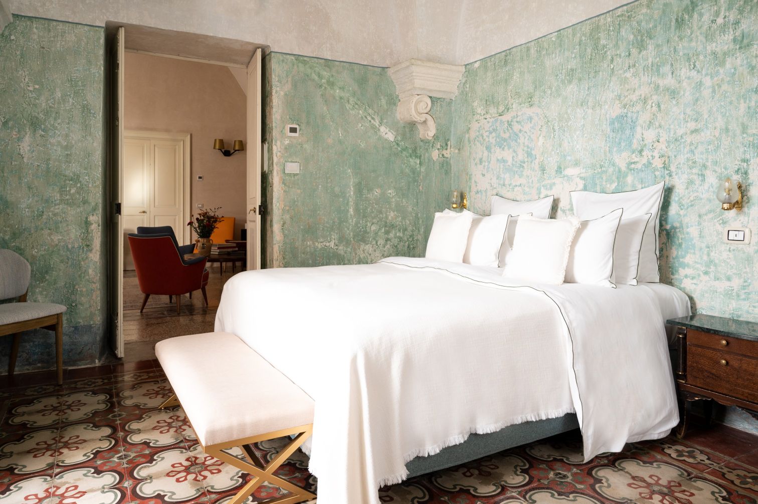 Cozy bedroom featuring a large bed with white linens, decorative pillows, and a stylish bench at the foot. The room showcases vintage green walls, intricate patterned tiled flooring, and a glimpse of a sitting area with colorful furniture in the background. Ideal for a relaxing stay. | Only on SopranoVillas