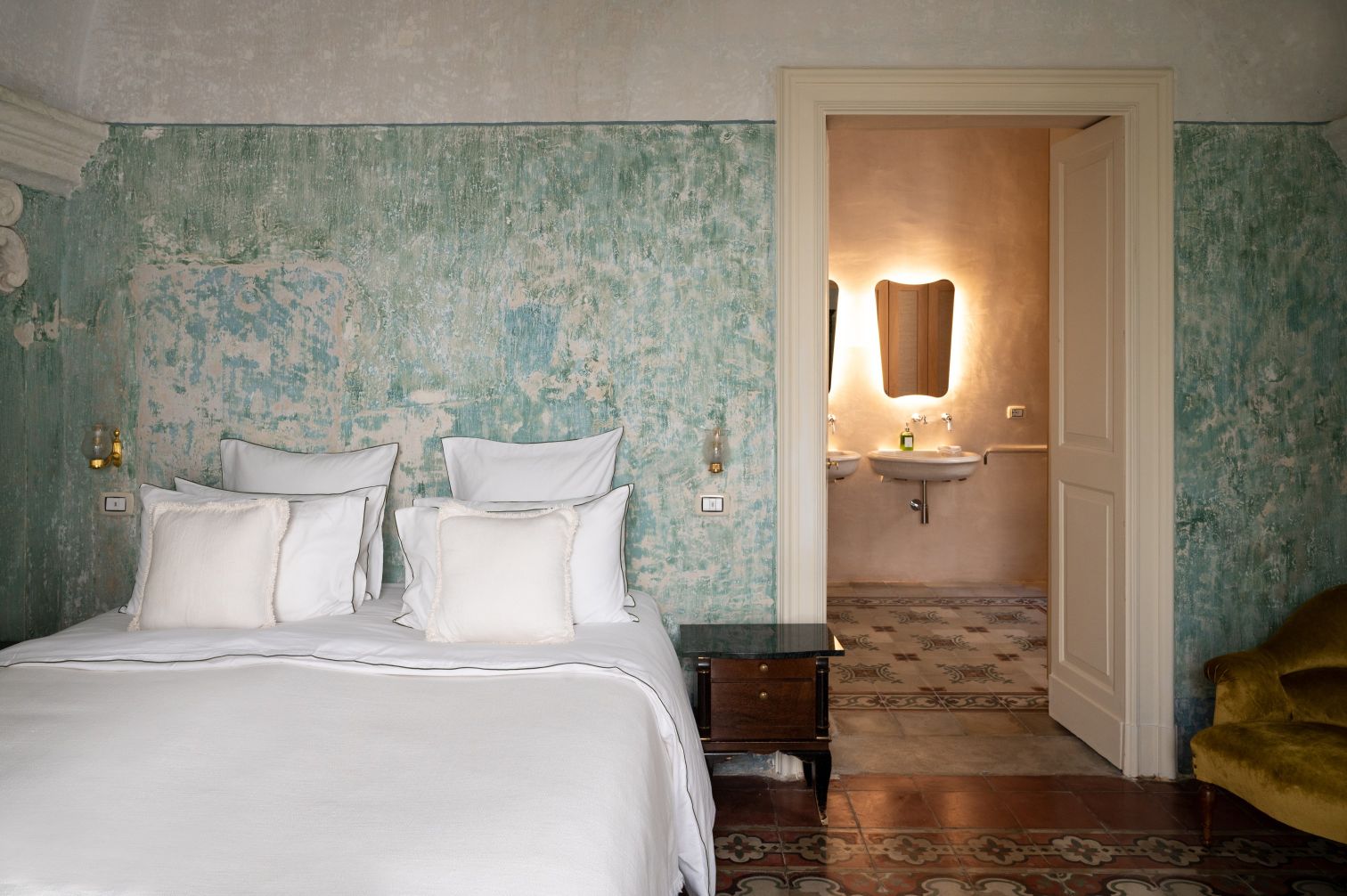 Cozy bedroom featuring a vintage design with a distressed green wall, plush bedding, and decorative pillows. The room opens to a stylish bathroom with modern fixtures and warm lighting. A small wooden nightstand complements the aesthetic, while a plush armchair adds comfort. | Only on SopranoVillas