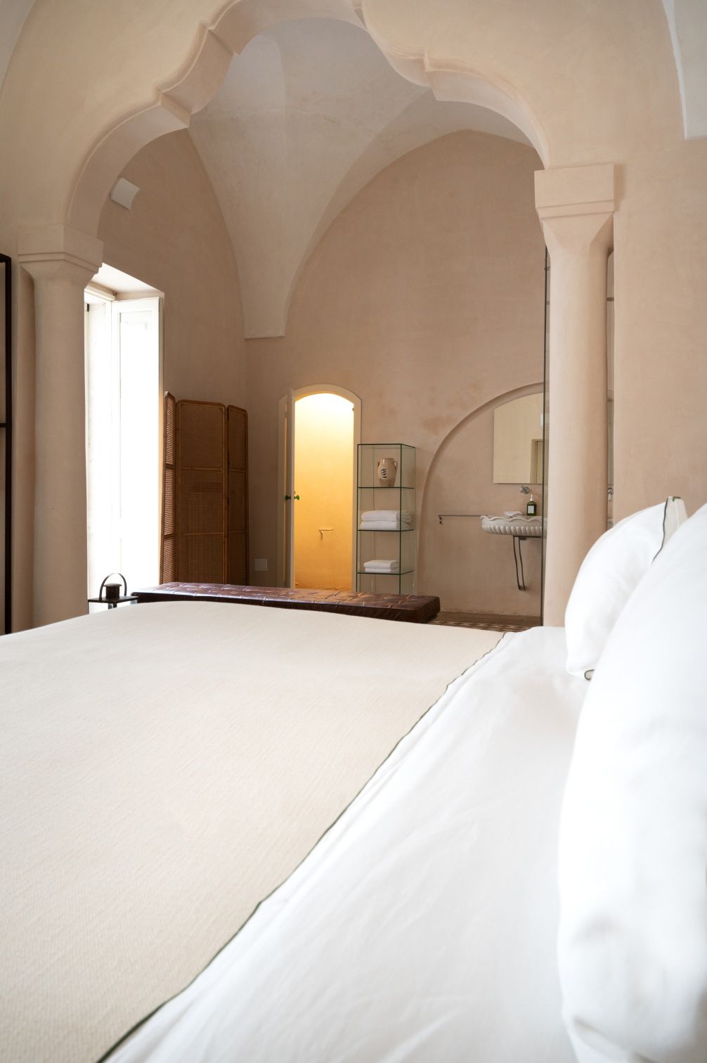 Spacious bedroom with arched ceilings featuring a neutral color palette, a large bed with white linens, and a wooden bench. The room includes a window with natural light, a decorative folding screen, and a modern bathroom area with a glass shelf and sink. | Only on SopranoVillas