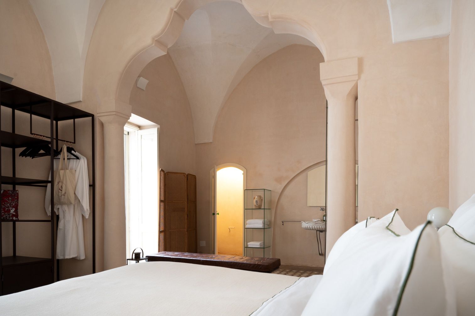 Elegant hotel room with arched ceilings, featuring a king-sized bed, modern furnishings, and a cozy seating area. The room includes a wardrobe with hanging clothes, a glass display for toiletries, and a bathroom entrance, all designed in soft, neutral tones for a serene atmosphere. | Only on SopranoVillas