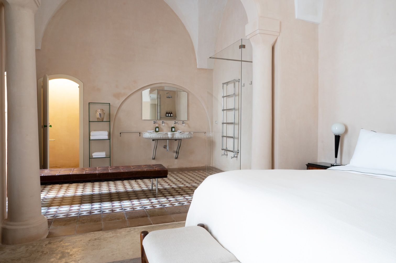 Stylish hotel room featuring a comfortable bed, a wooden bench, and an elegant bathroom area with modern fixtures and a unique tile floor design. | Only on SopranoVillas