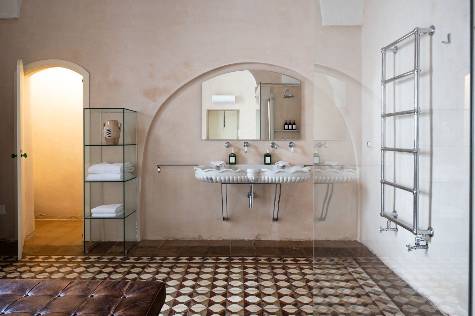 Modern bathroom interior featuring a unique shell-shaped sink, a glass shelf with neatly stacked towels, and stylish toiletries. The walls are adorned with a soft peach finish, while the floor showcases intricate patterned tiles. A sleek towel radiator and mirrors enhance the contemporary design. | Only on SopranoVillas