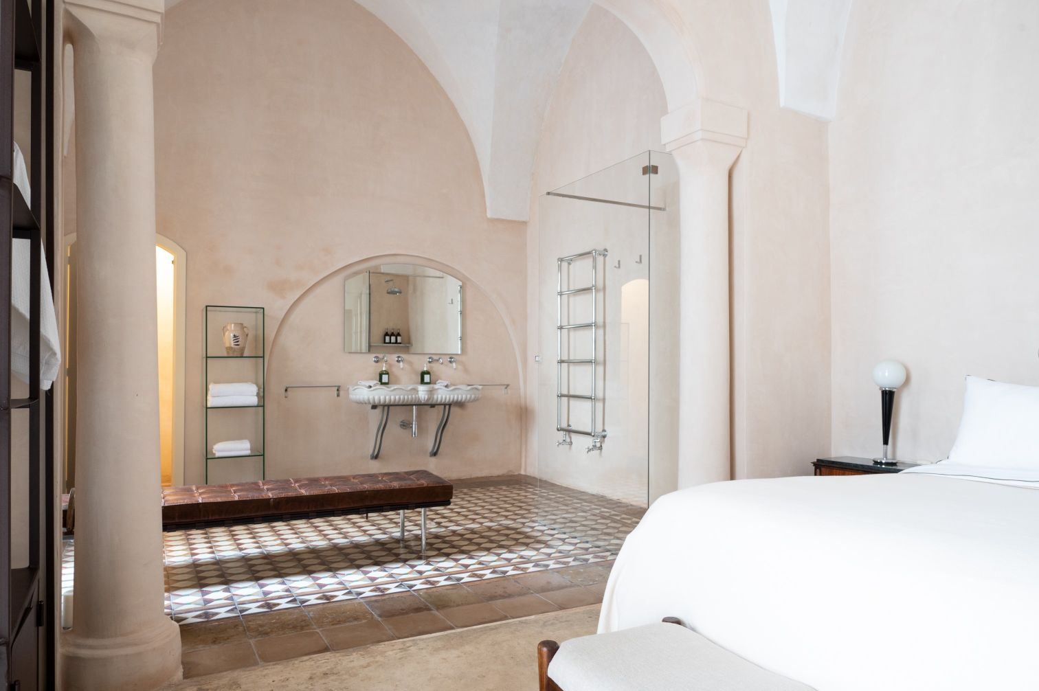 Spacious and elegantly designed bathroom featuring arched ceilings, a modern glass shower, and vintage-style sinks. The room includes a cozy bench, neatly arranged towels, and stylish decor elements, creating a serene atmosphere. | Only on SopranoVillas