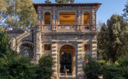 Historic stone building with intricate architectural details, featuring arched windows and a decorative facade, surrounded by lush greenery and flowering plants in a serene garden setting.