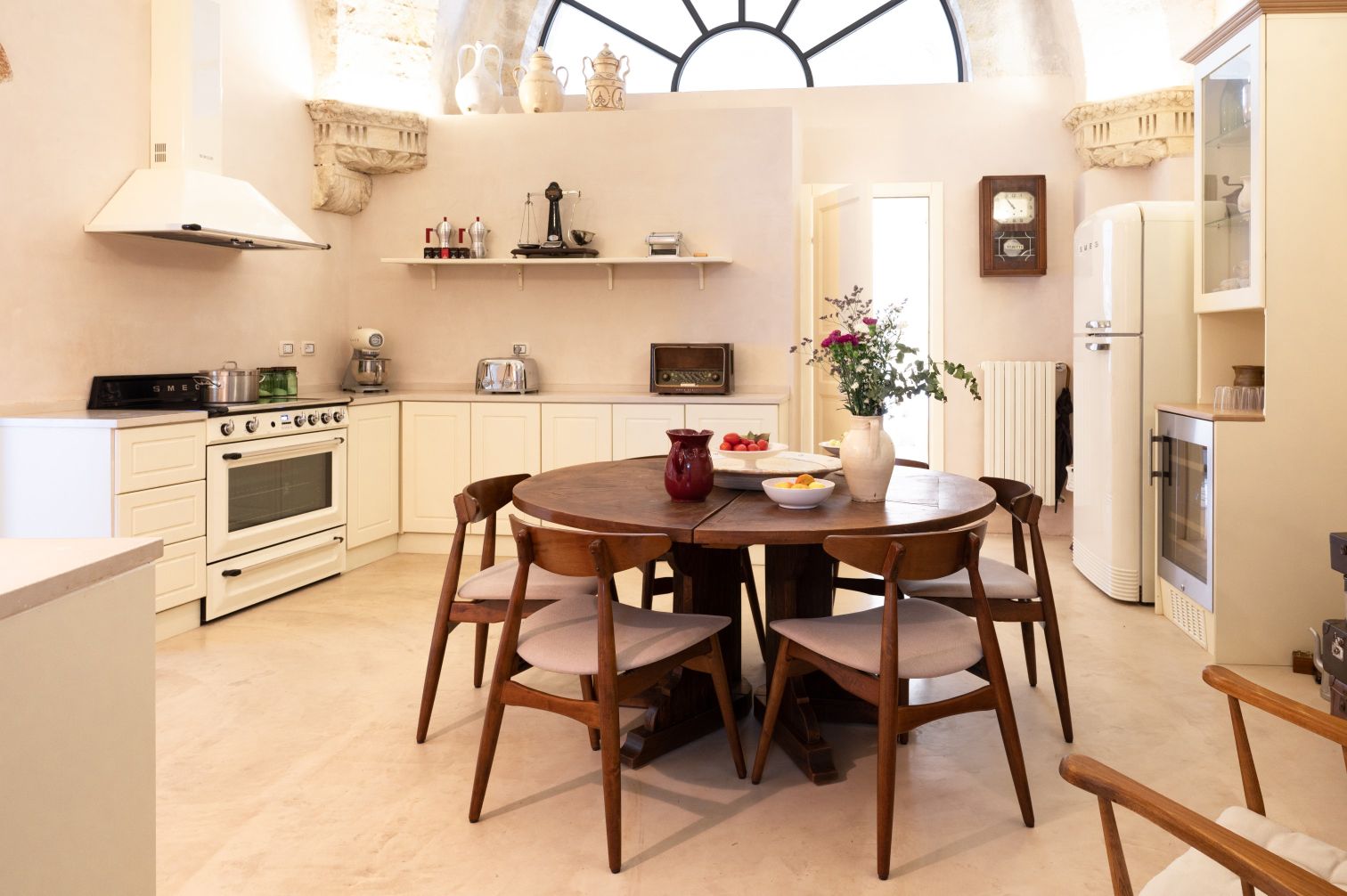 Modern kitchen interior featuring a round wooden dining table with six chairs, a white SMEG oven, and various kitchen appliances. The space is well-lit with natural light from a large window, and decorative elements include a vase of flowers and fruit bowls on the table. The cabinetry is in soft tones, complemented by a minimalist design. | Only on SopranoVillas