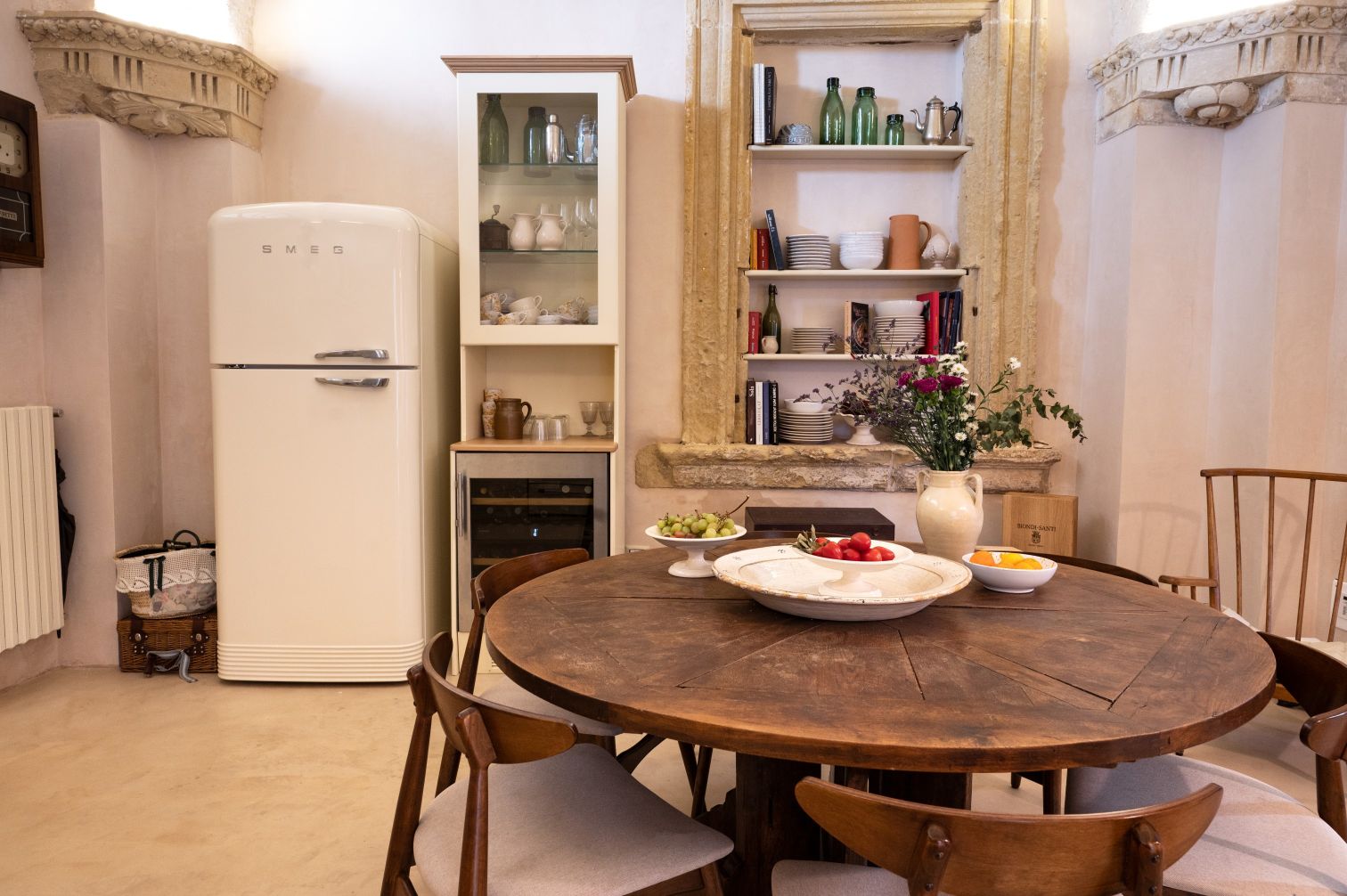 Bright and inviting kitchen interior featuring a retro-style SMEG refrigerator, wooden dining table with fresh fruit, and a display cabinet showcasing glassware and ceramics. The walls are adorned with rustic architectural details, creating a warm and cozy atmosphere. | Only on SopranoVillas