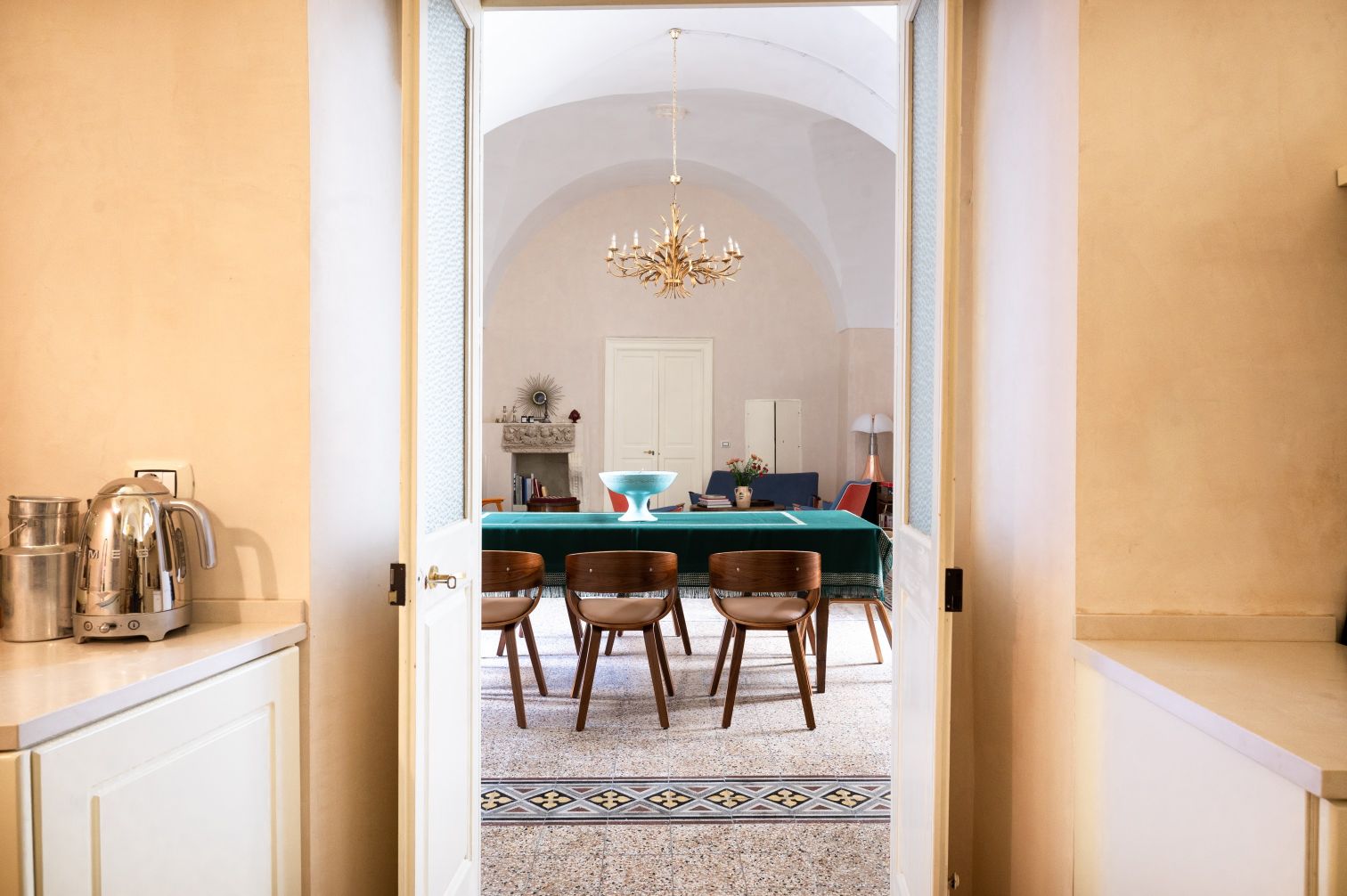 View from a kitchen doorway into a stylish dining area featuring a green table, wooden chairs, and elegant chandelier, with decorative elements and soft lighting creating a welcoming atmosphere. | Only on SopranoVillas