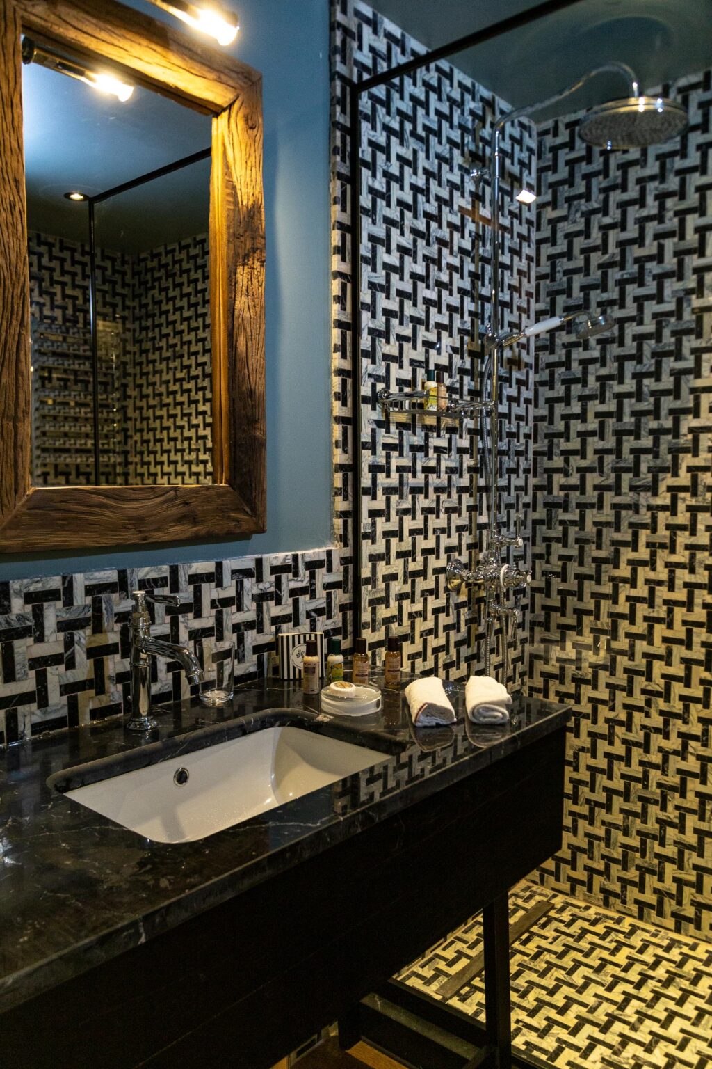 Modern bathroom featuring a sleek black marble sink, decorative patterned tiles, and a wooden-framed mirror. The shower area showcases a stylish chrome showerhead and an array of toiletries on the countertop, complemented by neatly folded towels. | Only on SopranoVillas