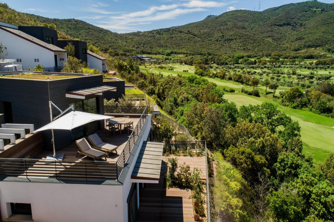 Modern hillside villa with a spacious wooden deck, featuring lounge chairs and a sunshade, overlooking lush green hills and a golf course. Ideal for relaxation and enjoying scenic views. | Only on SopranoVillas