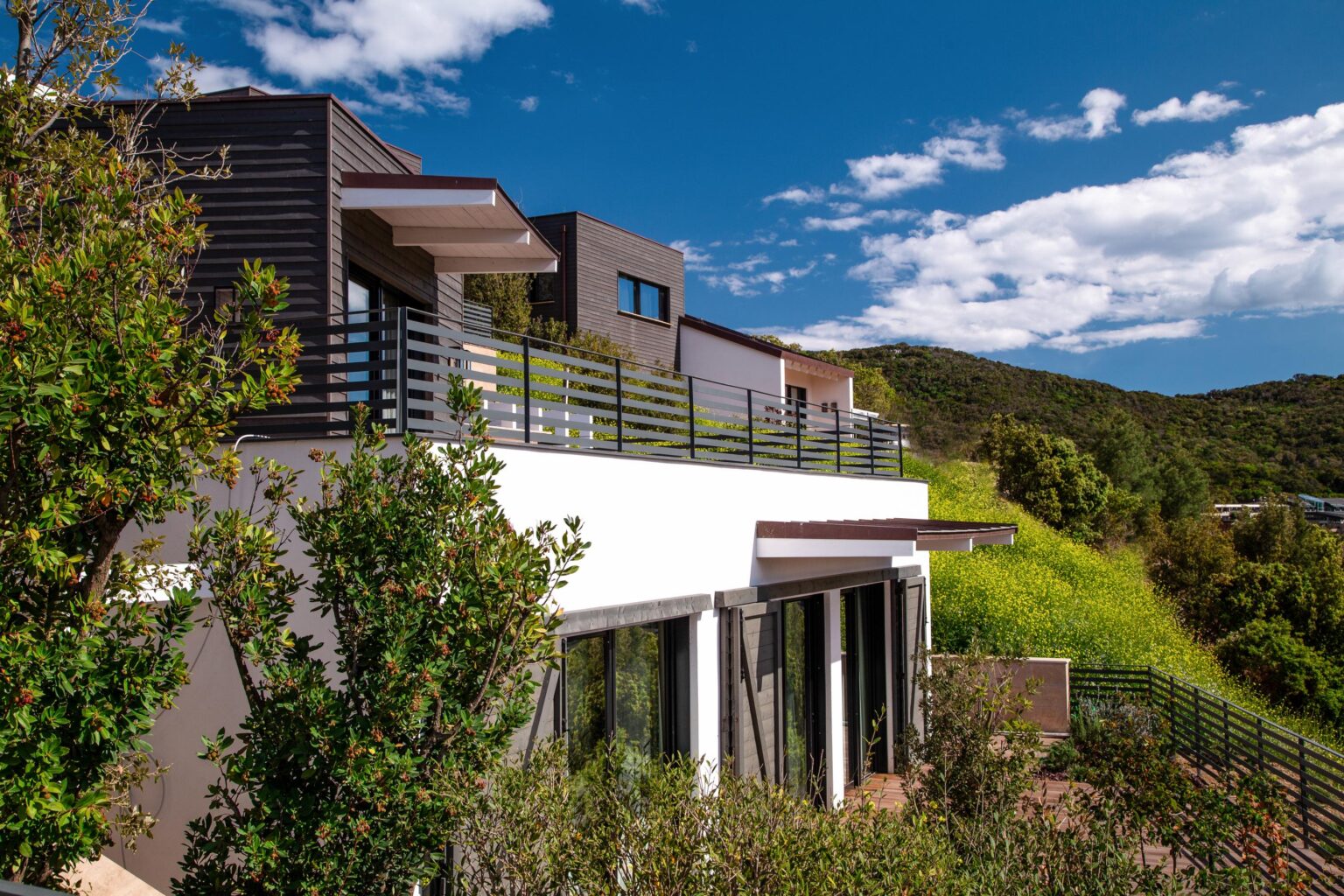 Modern hillside home with contemporary architecture, featuring expansive windows and a stylish balcony, surrounded by lush greenery and a clear blue sky. | Only on SopranoVillas