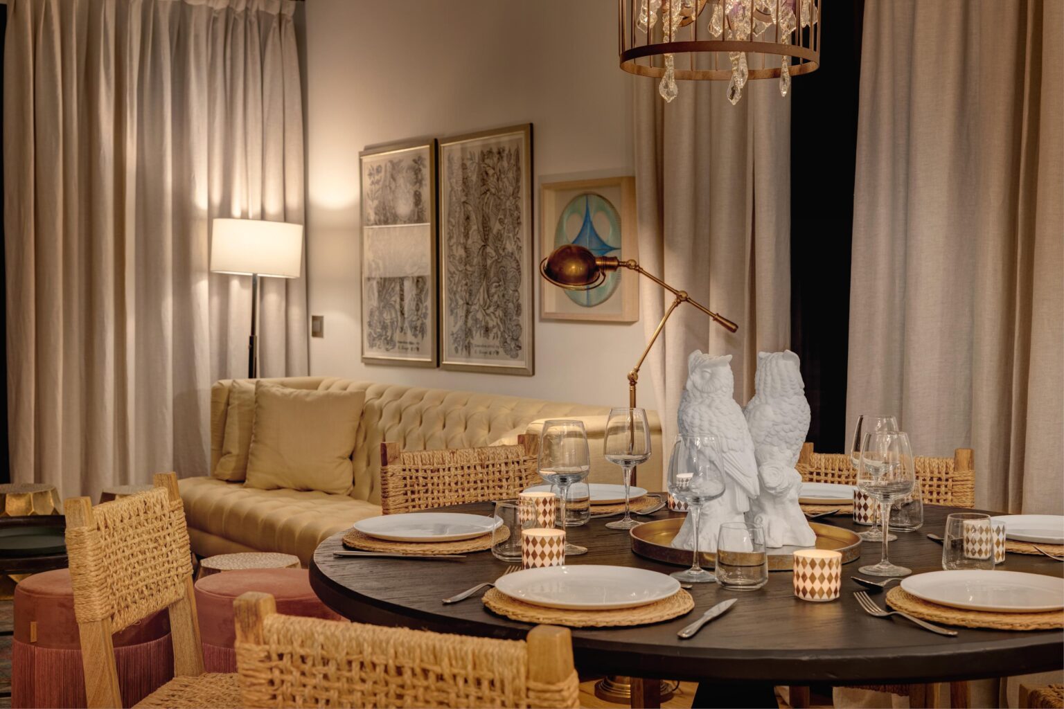 Elegant dining room featuring a round table set with white plates, glassware, and decorative accents. The space includes a cozy beige sofa, stylish artwork on the walls, and warm ambient lighting from a chandelier and a floor lamp. Textured curtains add depth to the decor, creating a sophisticated atmosphere. | Only on SopranoVillas